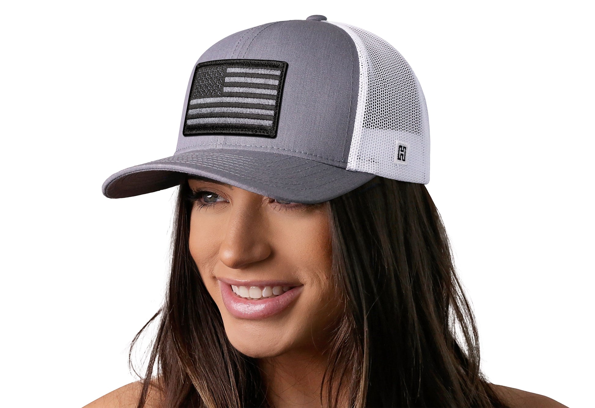 Gray/White trucker hat worn by a female model viewed from the left side and features a American Flag Embroidered Rectangle Patch displayed on the front panel. The hat has a classic mesh back design and a curved structured bill