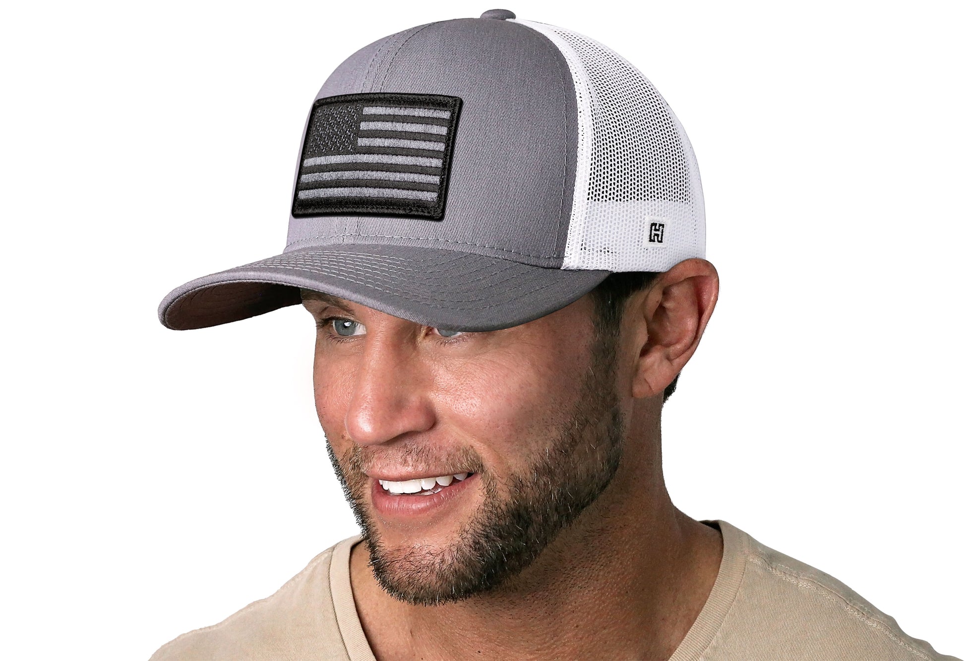 Gray/White trucker hat worn by a male model viewed from the left side and features a American Flag Embroidered Rectangle Patch displayed on the front panel. The hat has a classic mesh back design and a curved structured bill