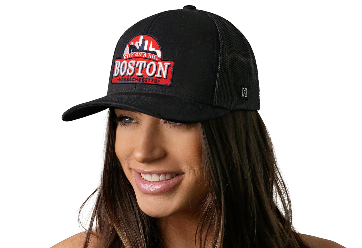 Black trucker hat worn by a female model viewed from the left side and features a Boston Cityscape Embroidered Patch displayed on the front panel. The hat has a classic mesh back design and a curved structured bill