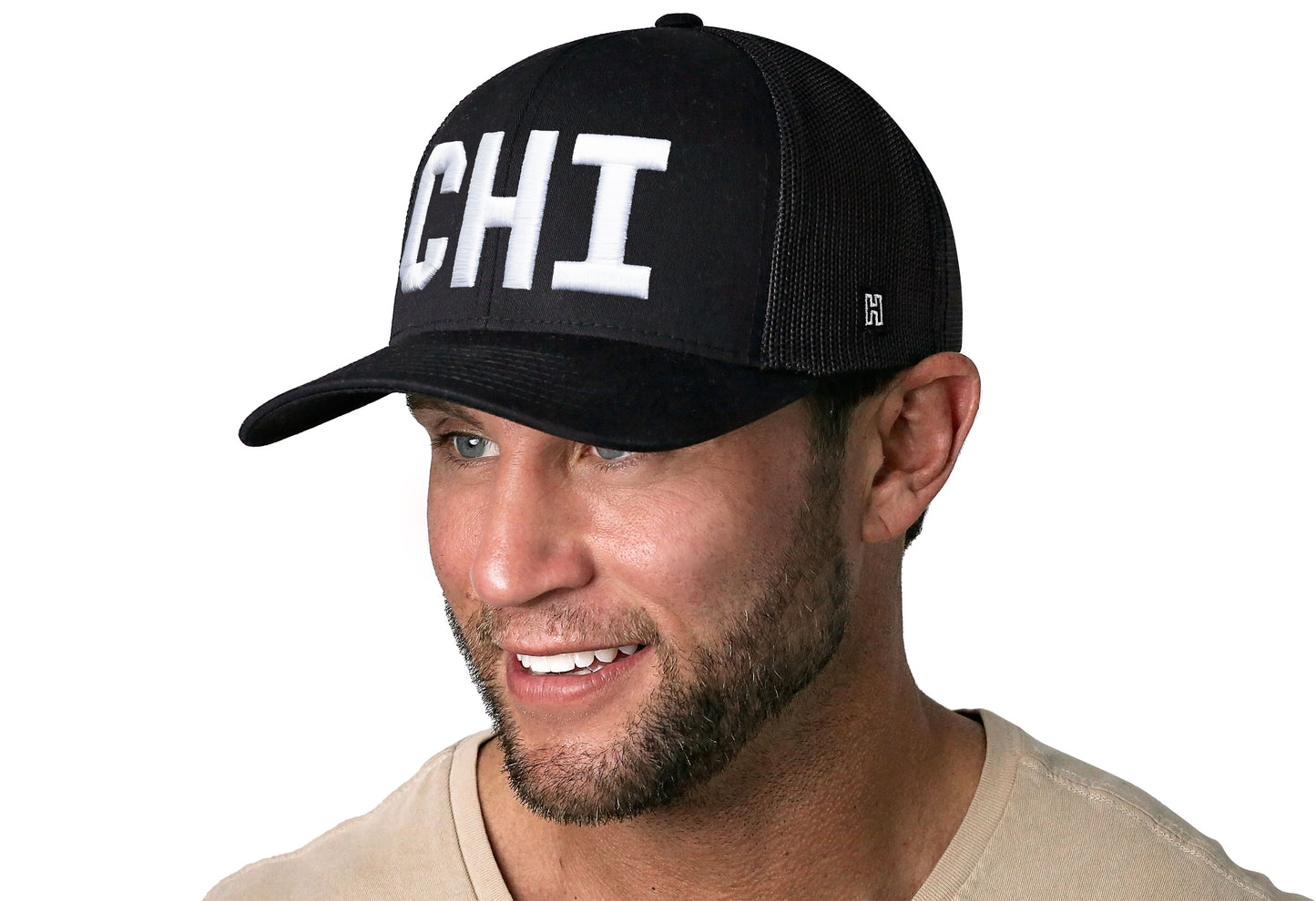 Black trucker hat worn by a male model viewed from the left side and features a White "CHI" Puff Embroidery Block displayed on the front panel. The hat has a classic mesh back design and a curved structured bill