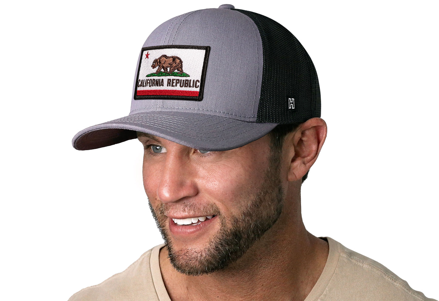 Gray-Black trucker hat worn by a male model viewed from the left side and features a California Flag Embroidered Rectangle Patch displayed on the front panel. The hat has a classic mesh back design and a curved structured bill
