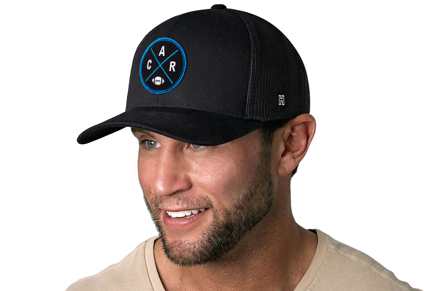 black trucker hat worn by a male model viewed from the left side and features a bold Carolina blue and black embroidered X patch displayed on the front panel. The hat has a classic mesh back design and a curved structured bill