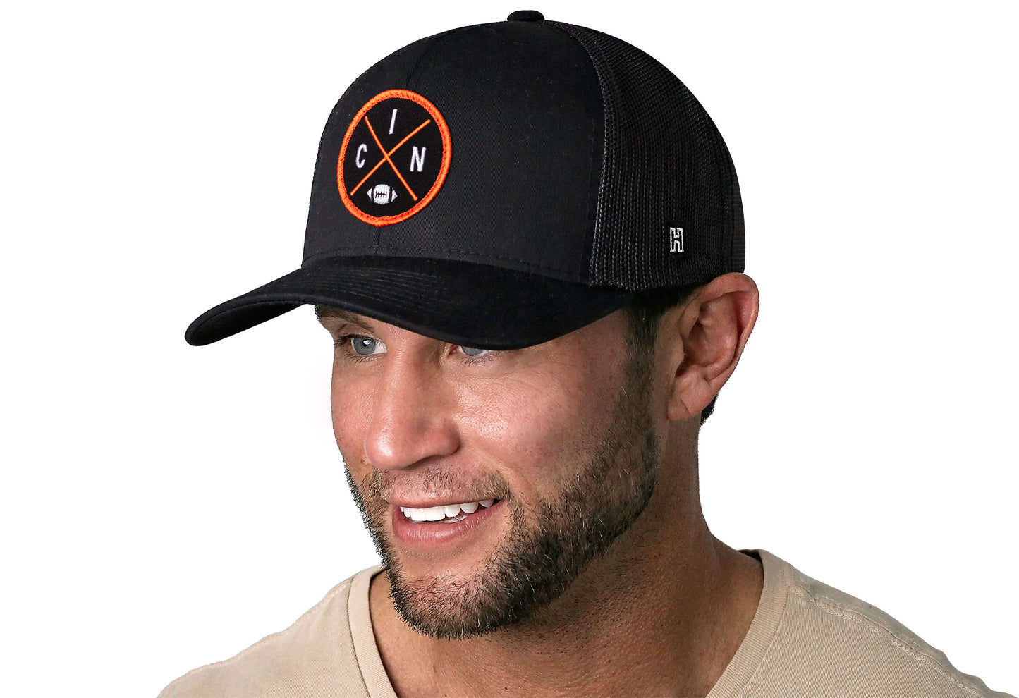 black trucker hat worn by a male model viewed from the left side and features a bold orange and black embroidered X patch displayed on the front panel. The hat has a classic mesh back design and a curved structured bill