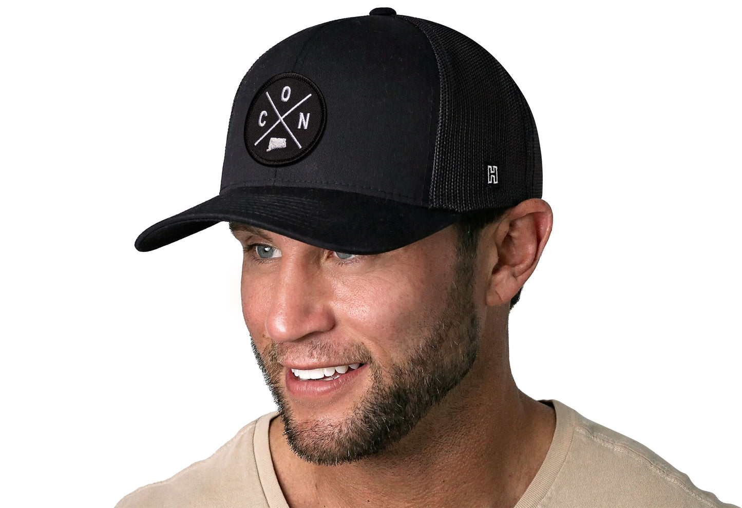 Black trucker hat worn by a male model viewed from the left side and features a Connecticut CON X Embroidered Circle Patch displayed on the front panel. The hat has a classic mesh back design and a curved structured bill