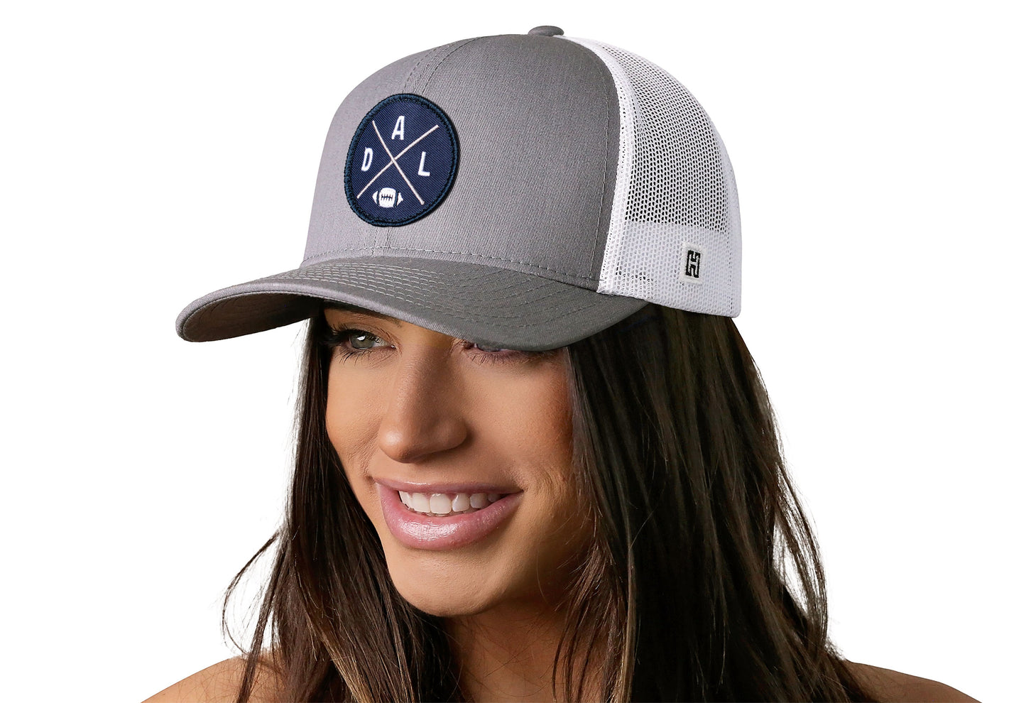 gray-white trucker hat worn by a female model viewed from the left side and features a bold navy, silver, and white embroidered X patch displayed on the front panel. The hat has a classic mesh back design and a curved structured bill
