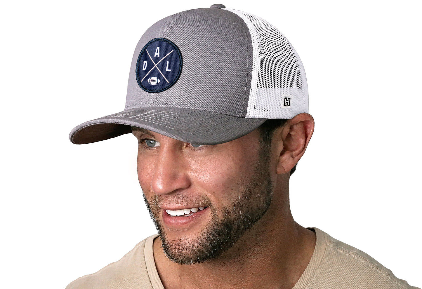 gray-white trucker hat worn by a male model viewed from the left side and features a bold navy, silver, and white embroidered X patch displayed on the front panel. The hat has a classic mesh back design and a curved structured bill