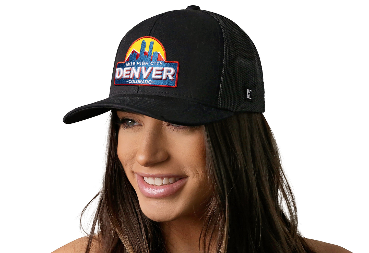Black trucker hat worn by a female model viewed from the left side and features a Denver Cityscape Embroidered Patch displayed on the front panel. The hat has a classic mesh back design and a curved structured bill