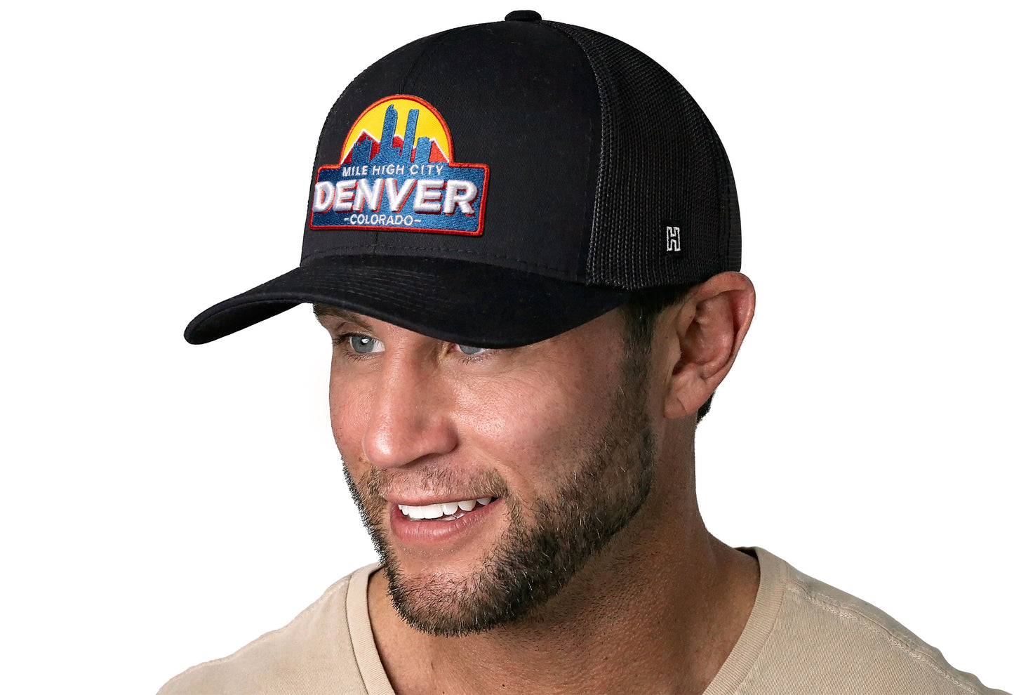 Black trucker hat worn by a male model viewed from the left side and features a Denver Cityscape Embroidered Patch displayed on the front panel. The hat has a classic mesh back design and a curved structured bill