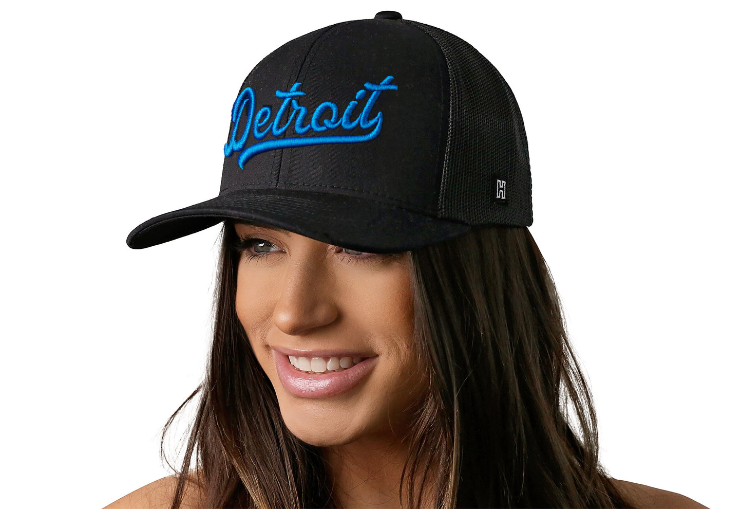 black trucker hat worn by a female model viewed from the left side and features a Blue "Detroit" Puff Script 3D Embroidery displayed on the front panel. The hat has a classic mesh back design and a curved structured bill