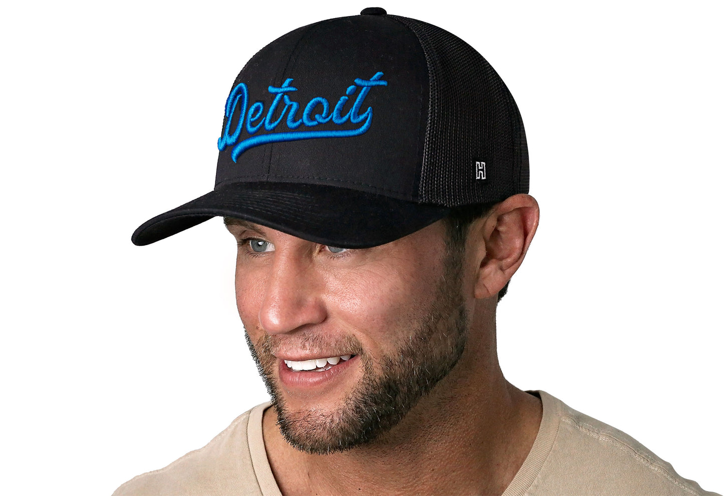 black trucker hat worn by a male model viewed from the left side and features a Blue "Detroit" Puff Script 3D Embroidery displayed on the front panel. The hat has a classic mesh back design and a curved structured bill