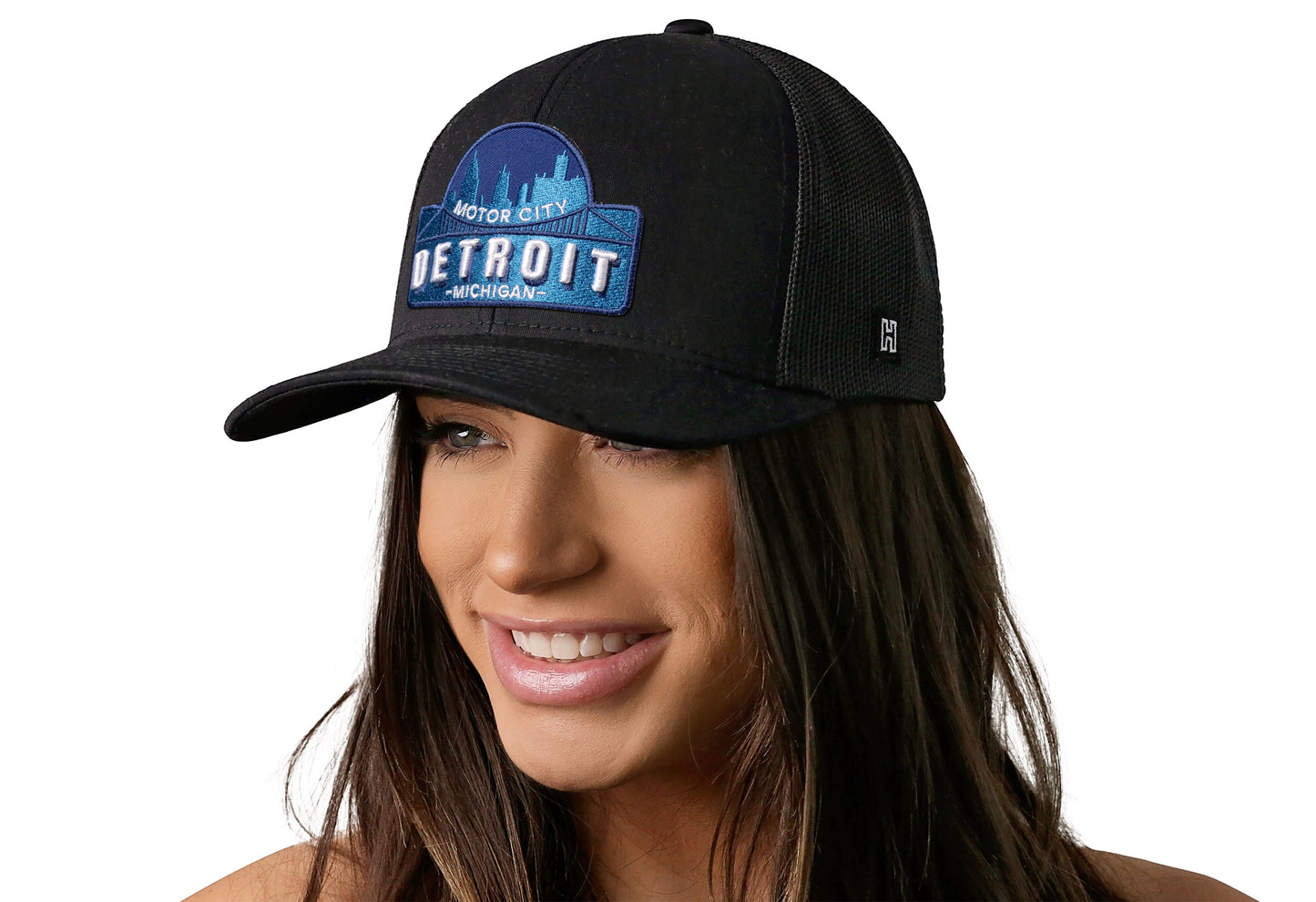 Black trucker hat worn by a female model viewed from the left side and features a Detroit Cityscape Embroidered Patch displayed on the front panel. The hat has a classic mesh back design and a curved structured bill