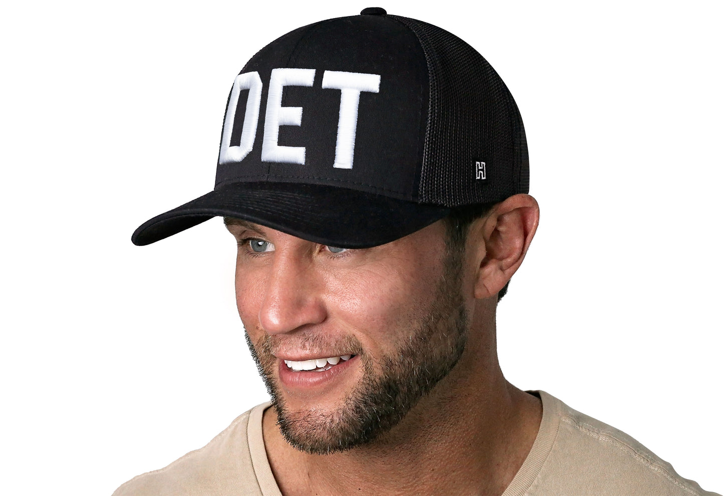 Black trucker hat worn by a male model viewed from the left side and features a DET 3-D Puff Direct Embroidery displayed on the front panel. The hat has a classic mesh back design and a curved structured bill