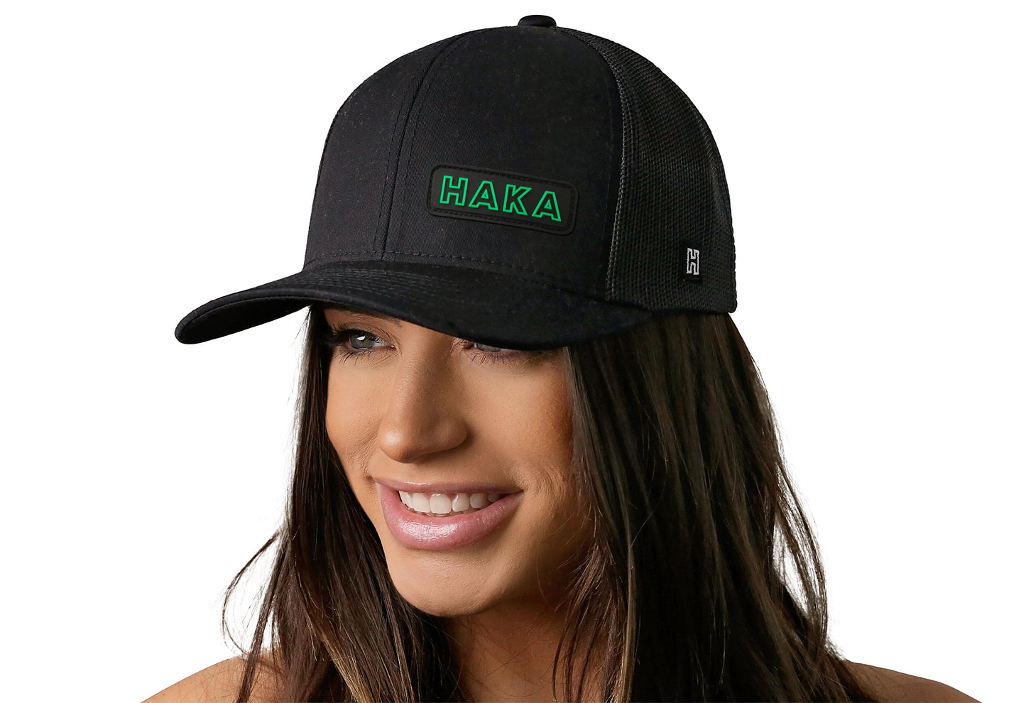Black trucker hat worn by a female model viewed from the left side and features a HAKA Rectangle Rubber Patch displayed on the front panel. The hat has a classic mesh back design and a curved structured bill
