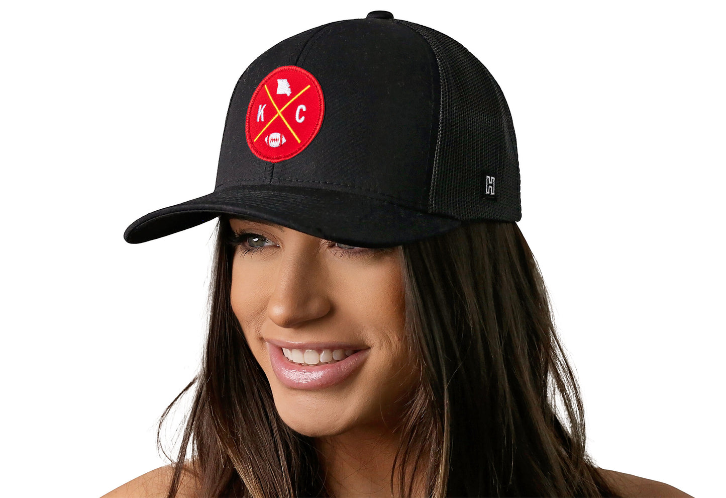black trucker hat worn by a female model viewed from the left side and features a bold red and gold embroidered X patch displayed on the front panel. The hat has a classic mesh back design and a curved structured bill