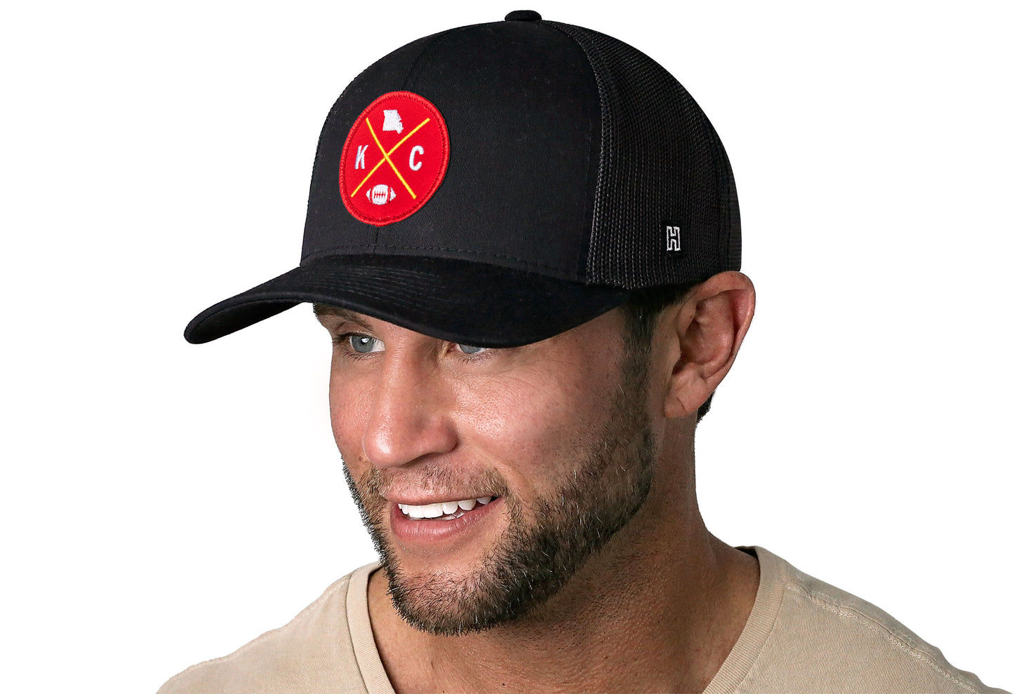 black trucker hat worn by a male model viewed from the left side and features a bold red and gold embroidered X patch displayed on the front panel. The hat has a classic mesh back design and a curved structured bill