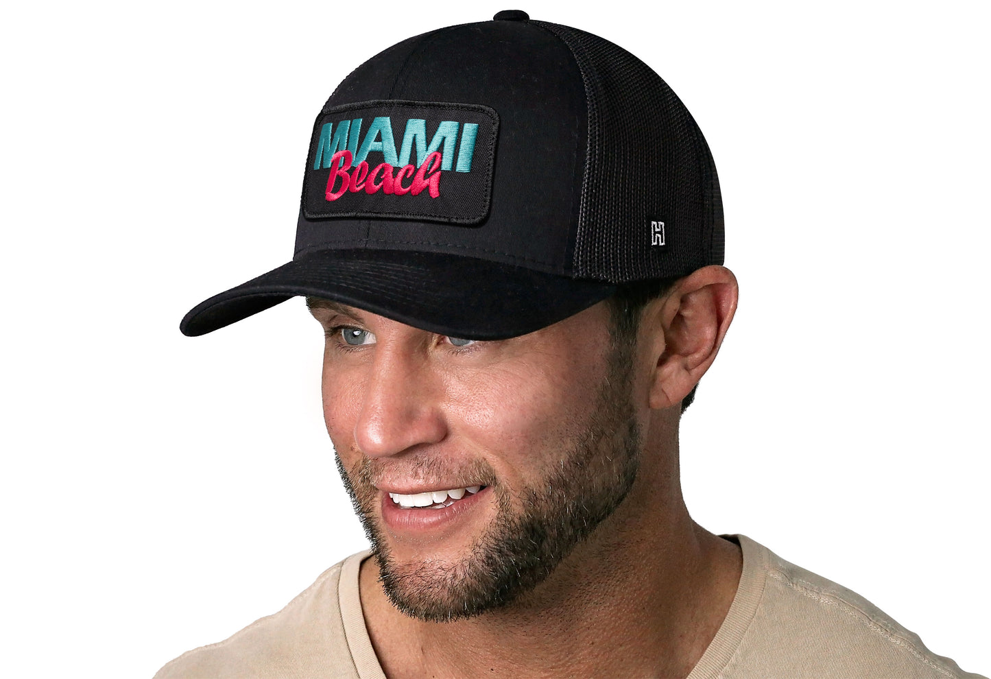 Black trucker hat worn by a male model viewed from the left side and features a Miami Beach Embroidered Circle Patch displayed on the front panel. The hat has a classic mesh back design and a curved structured bill