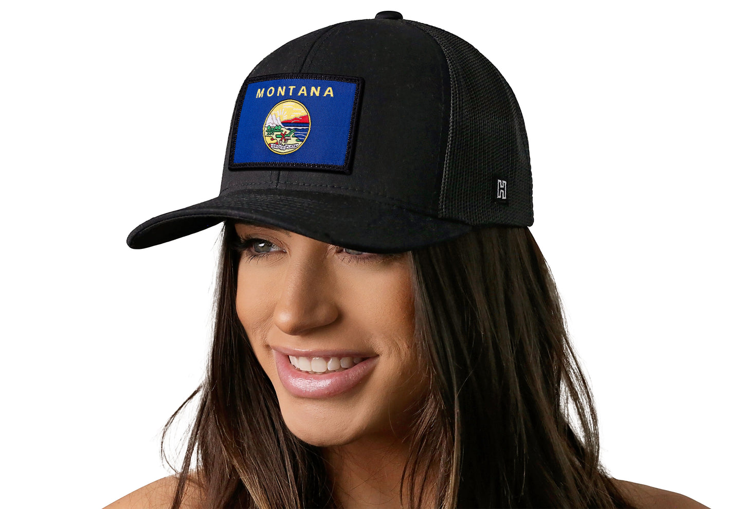 Black trucker hat worn by a female model viewed from the left side and features a Montana Flag Woven Rectangle Patch displayed on the front panel. The hat has a classic mesh back design and a curved structured bill