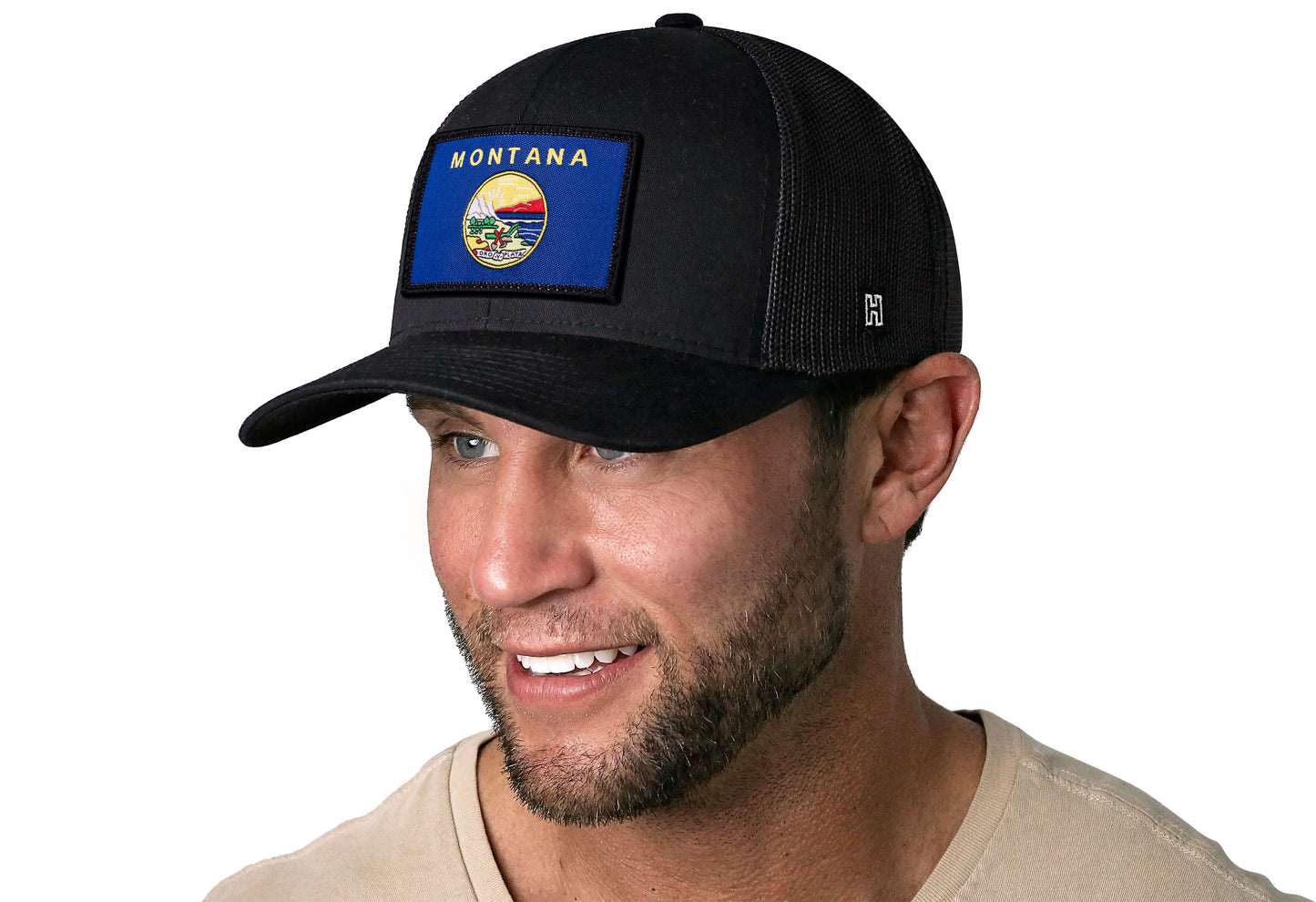 Black trucker hat worn by a male model viewed from the left side and features a Montana Flag Woven Rectangle Patch displayed on the front panel. The hat has a classic mesh back design and a curved structured bill