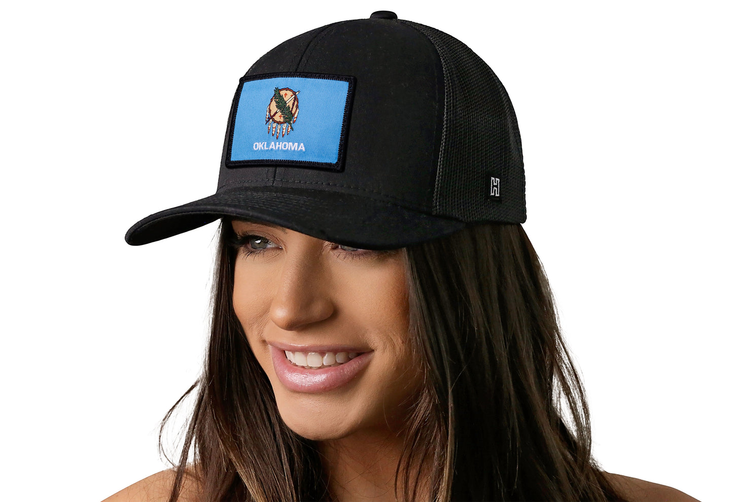 Black trucker hat worn by a female model viewed from the left side and features a Oklahoma Flag Woven Rectangule Patch displayed on the front panel. The hat has a classic mesh back design and a curved structured bill