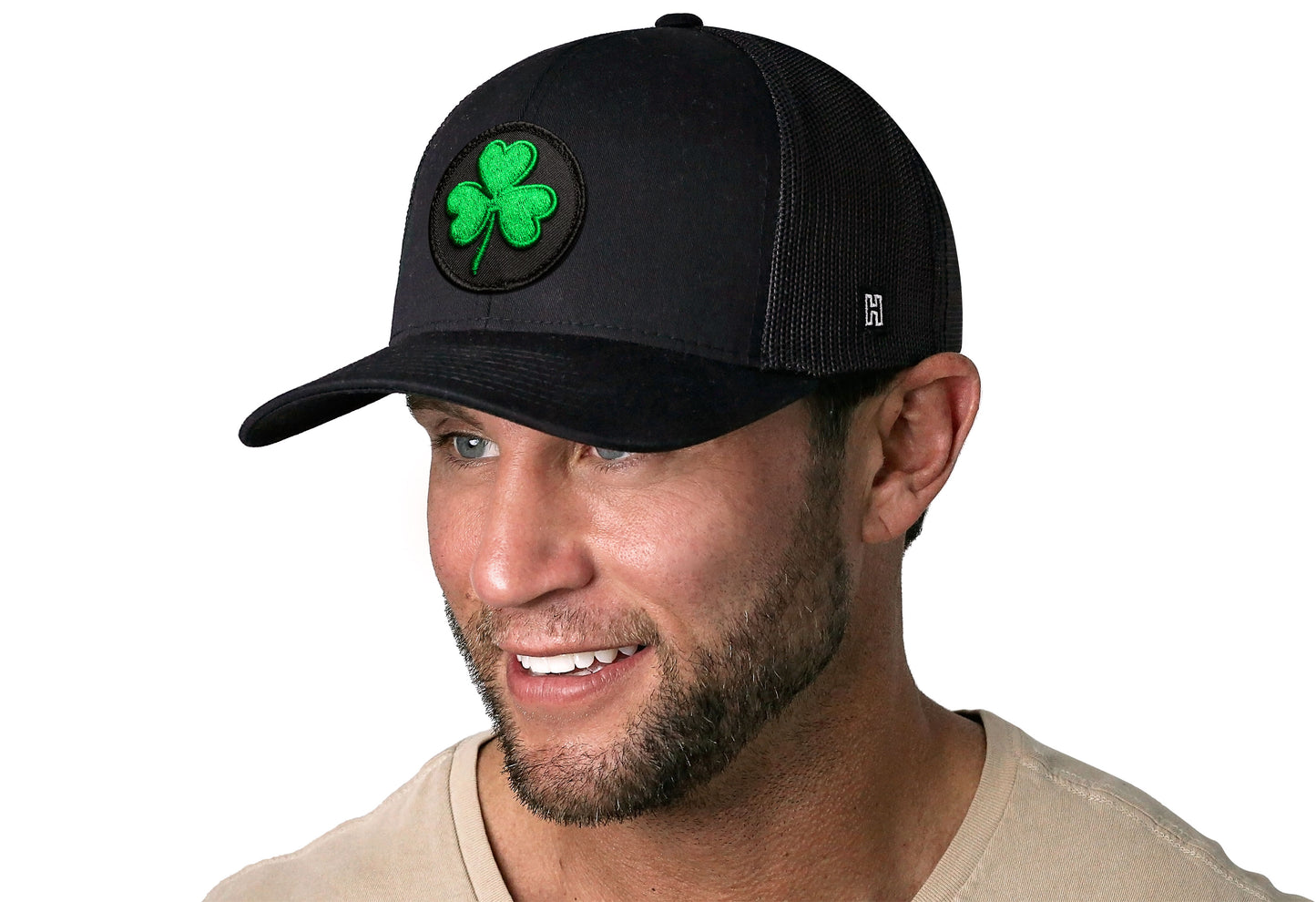 Black trucker hat worn by a male model viewed from the left side and features a Shamrock Black St Patrick's Day Embroidered Patch displayed on the front panel. The hat has a classic mesh back design and a curved structured bill