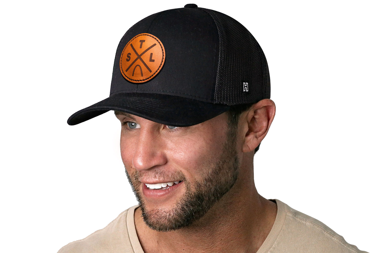 Black trucker hat worn by a male model viewed from the left side and features a St Louis STL X Leather Circle Patch displayed on the front panel. The hat has a classic mesh back design and a curved structured bill