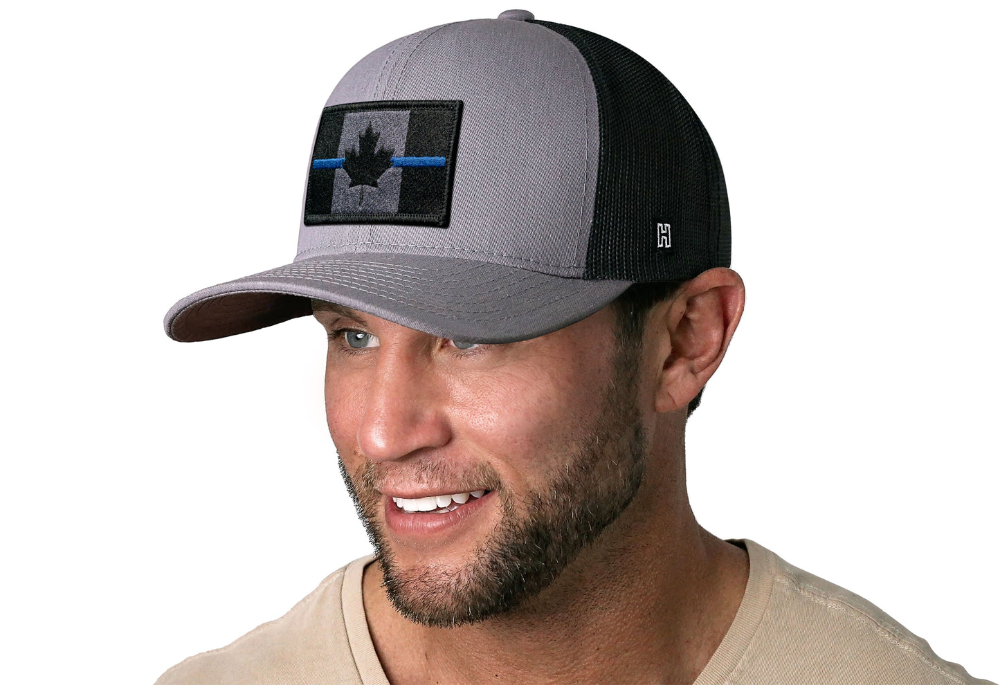 Gray-Black trucker hat worn by a male model viewed from the left side and features a Thin Blue Canadian Flag Embroidered Rectangle Patch displayed on the front panel. The hat has a classic mesh back design and a curved structured bill
