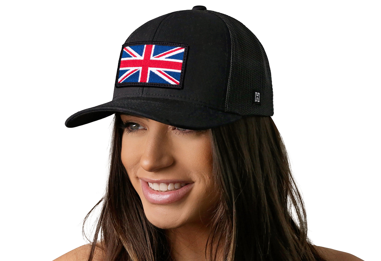 Black trucker hat worn by a female model viewed from the left side and features a United Kingdom Flag Embroidered Rectangle Patch displayed on the front panel. The hat has a classic mesh back design and a curved structured bill
