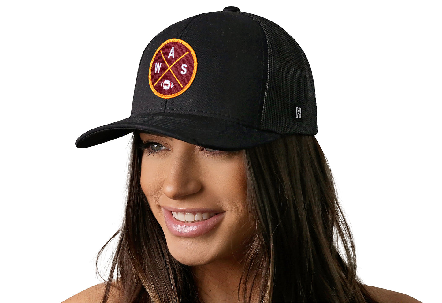 black trucker hat worn by a female model viewed from the left side and features a bold burgundy and gold embroidered X patch displayed on the front panel. The hat has a classic mesh back design and a curved structured bill
