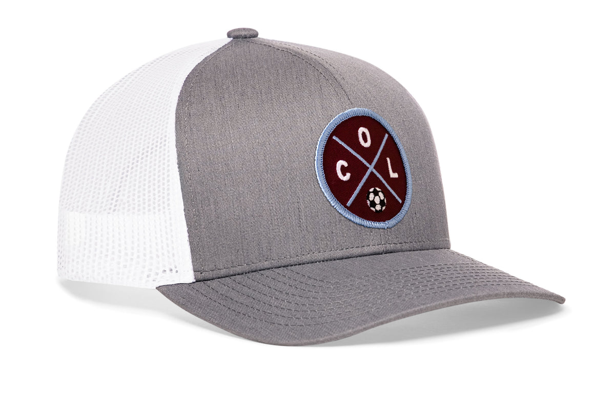 gray-white trucker hat viewed from the right side, showcasing a bold burgundy and blue embroidered X patch prominently displayed on the front