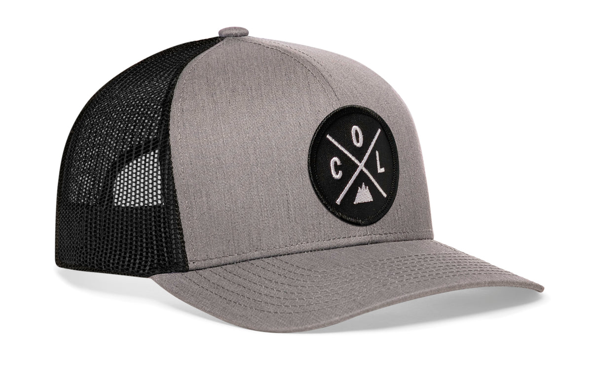 gray-black trucker hat viewed from the right side, showcasing a bold black and white embroidered X patch prominently displayed on the front