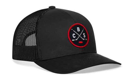 black trucker hat viewed from the right side, showcasing a navy, red and gray hockey embroidered X patch prominently displayed on the front