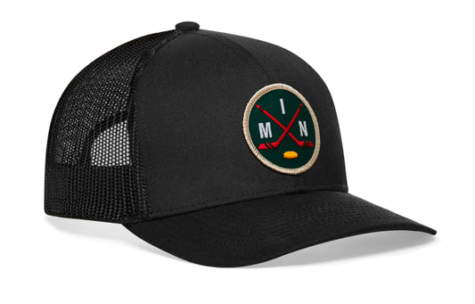 black trucker hat viewed from the right side, showcasing a green, red and gold hockey embroidered X patch prominently displayed on the front