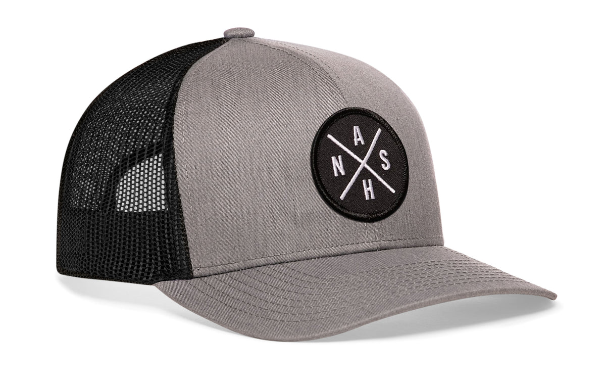 gray-black trucker hat viewed from the right side, showcasing a bold black and white embroidered X patch prominently displayed on the front