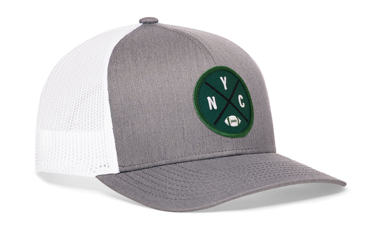 gray-white trucker hat viewed from the right side, showcasing a bold green, white and black embroidered X patch prominently displayed on the front