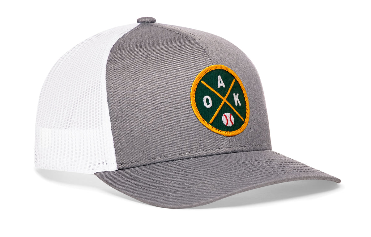 gray-white trucker hat viewed from the right side, showcasing a bold green and gold embroidered X patch prominently displayed on the front