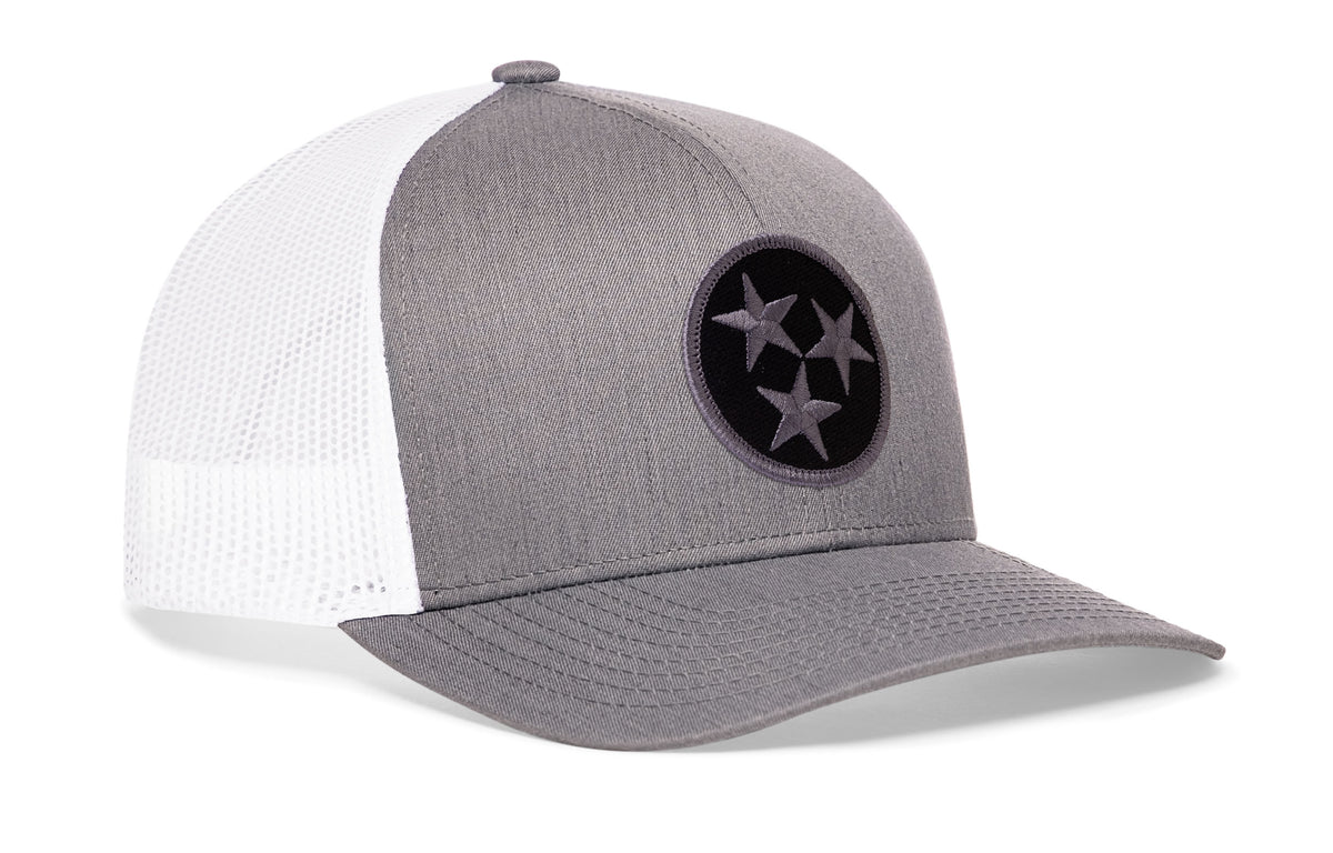 gray-white trucker hat viewed from the right side, showcasing a bold black and gray embroidered flag patch prominently displayed on the front