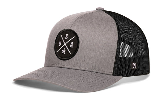 USA X Trucker Hat, gray-black snapback showing Haka brandmark on left side of cap.