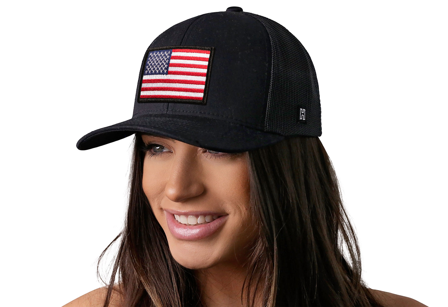 black trucker hat worn by a female model viewed from the left side and features a American Flag Embroidered Rectangle Patch displayed on the front panel. The hat has a classic mesh back design and a curved structured bill
