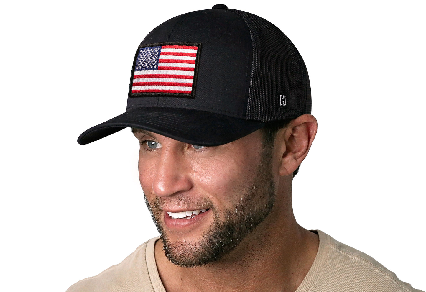 black trucker hat worn by a male model viewed from the left side and features a American Flag Embroidered Rectangle Patch displayed on the front panel. The hat has a classic mesh back design and a curved structured bill