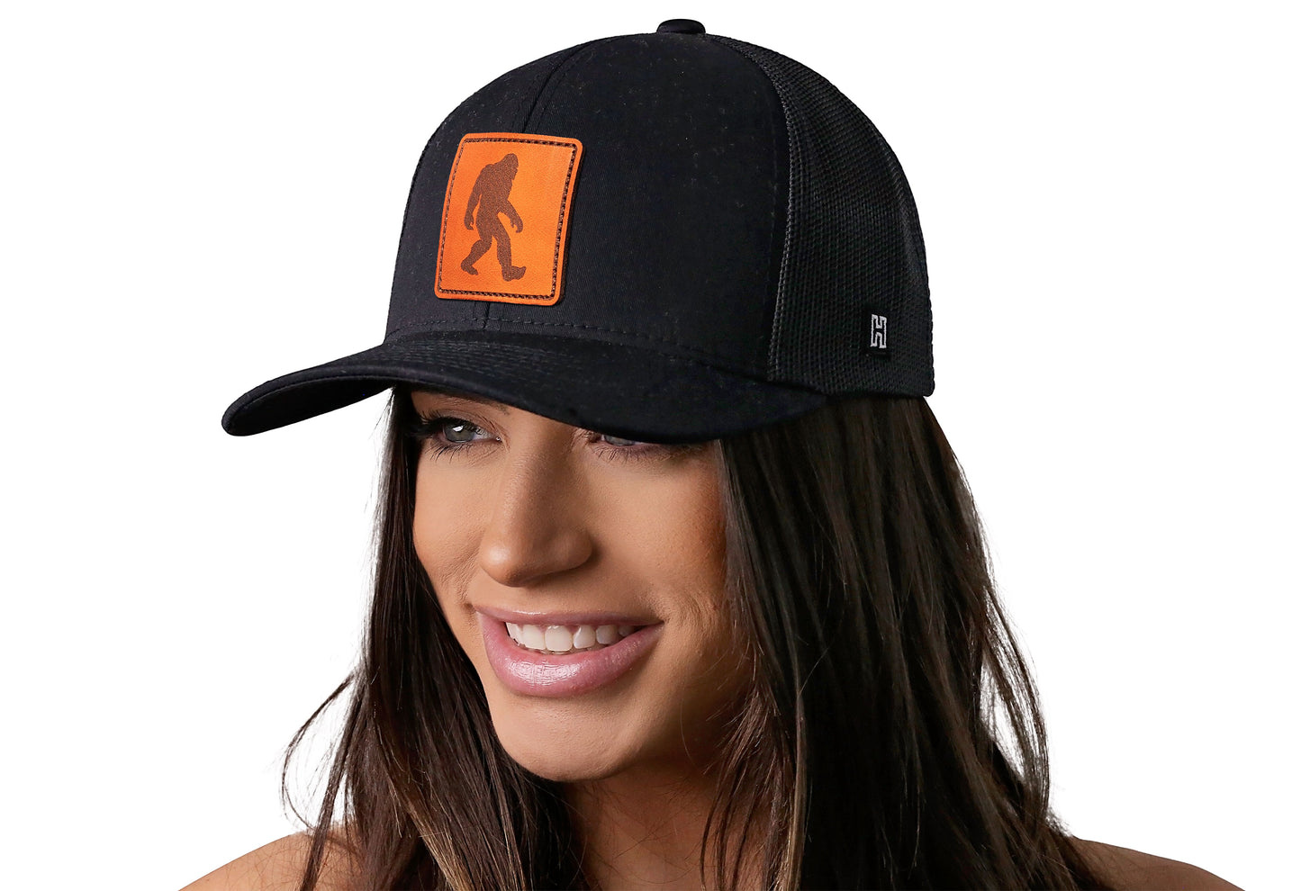 Black trucker hat worn by a female model viewed from the left side and features a Bigfoot Leather Black Sasquatch Leather displayed on the front panel. The hat has a classic mesh back design and a curved structured bill