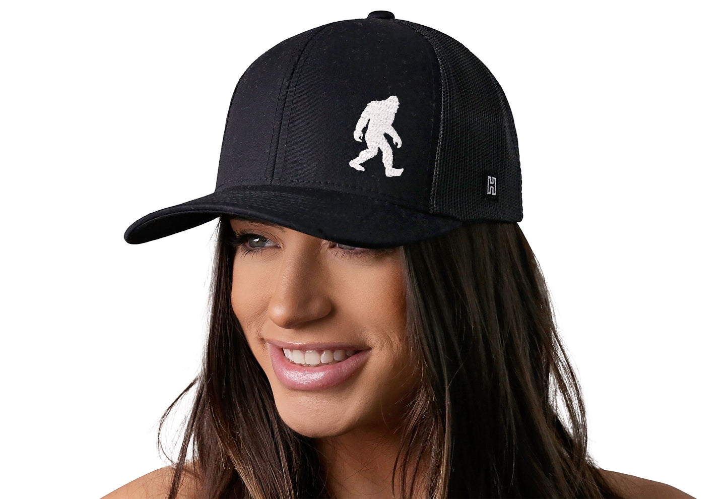Black trucker hat worn by a female model viewed from the left side and features a Bigfoot Black Sasquatch Flat Embroidery displayed on the front panel. The hat has a classic mesh back design and a curved structured bill