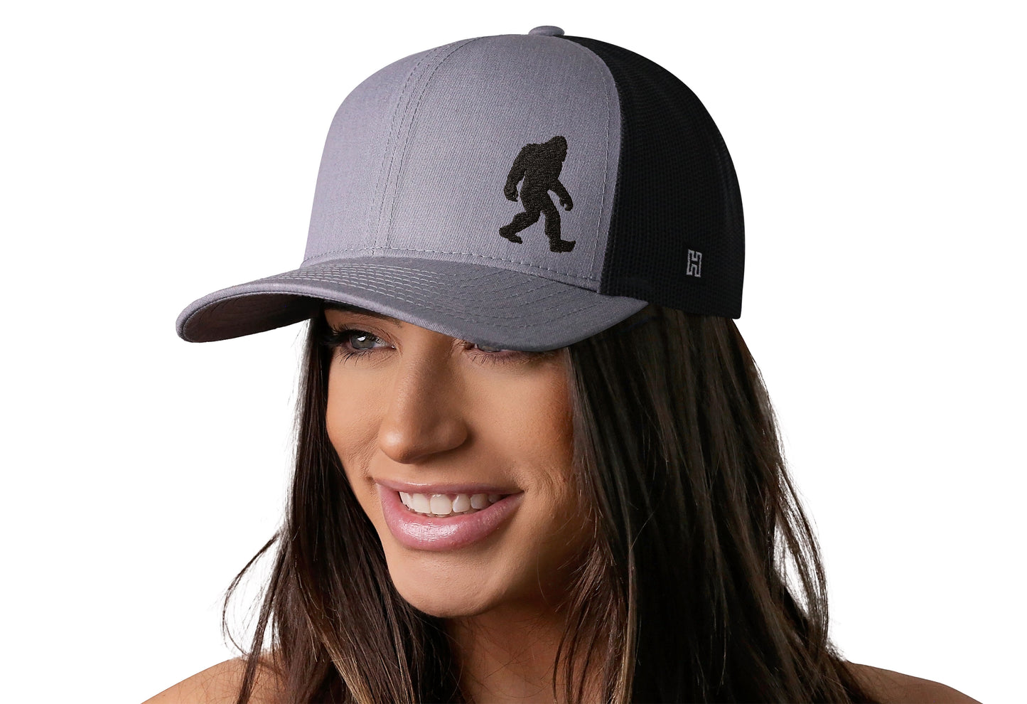 Gray-Black trucker hat worn by a female model viewed from the left side and features a Bigfoot Gray-Black Sasquatch Flat Embroidery displayed on the front panel. The hat has a classic mesh back design and a curved structured bill