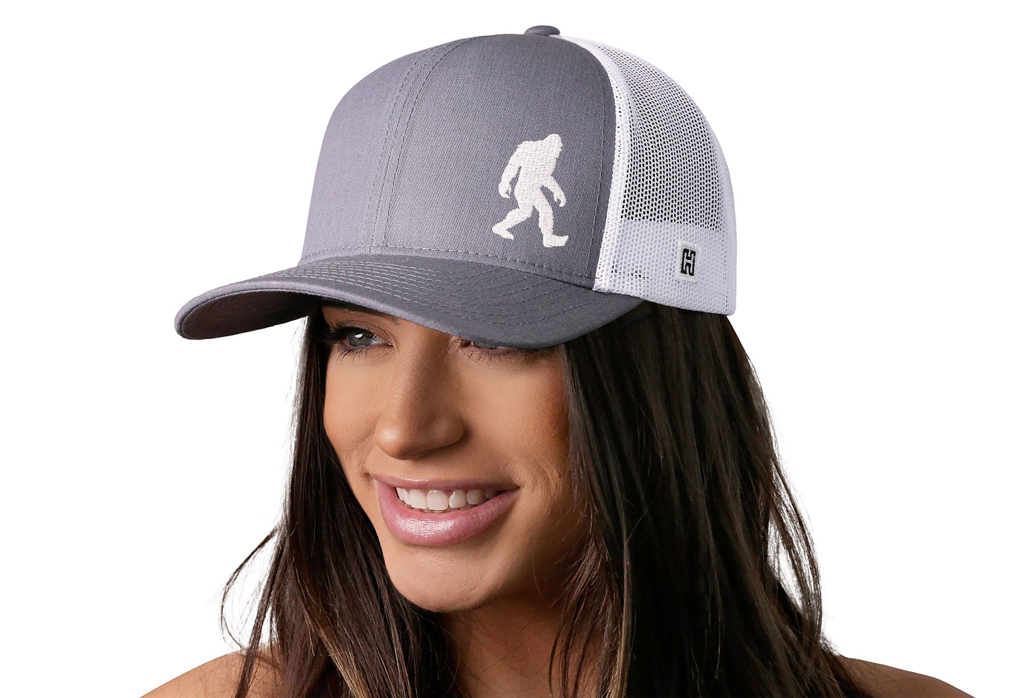 Gray-White trucker hat worn by a female model viewed from the left side and features a Bigfoot Sasquatch Flat Embroidery displayed on the front panel. The hat has a classic mesh back design and a curved structured bill