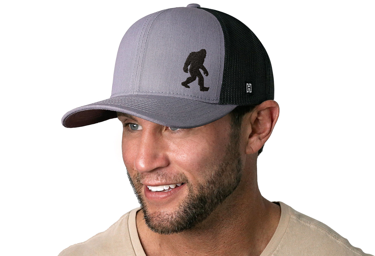 Gray-Black trucker hat worn by a male model viewed from the left side and features a Bigfoot Gray-Black Sasquatch Flat Embroidery displayed on the front panel. The hat has a classic mesh back design and a curved structured bill