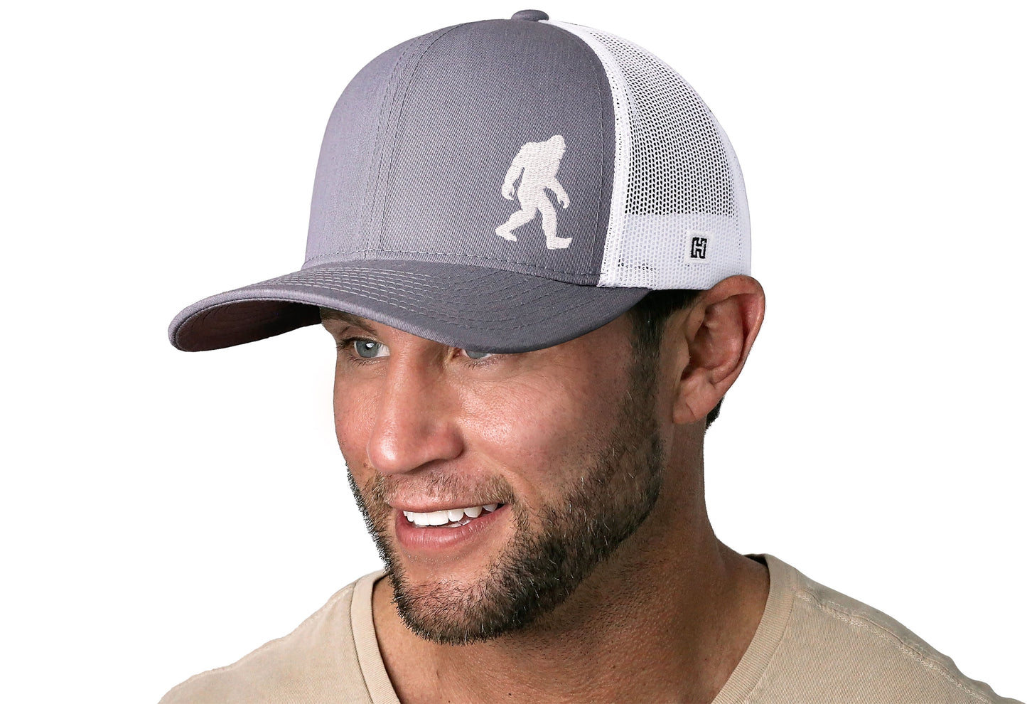 Gray-White trucker hat worn by a male model viewed from the left side and features a Bigfoot Sasquatch Flat Embroidery displayed on the front panel. The hat has a classic mesh back design and a curved structured bill