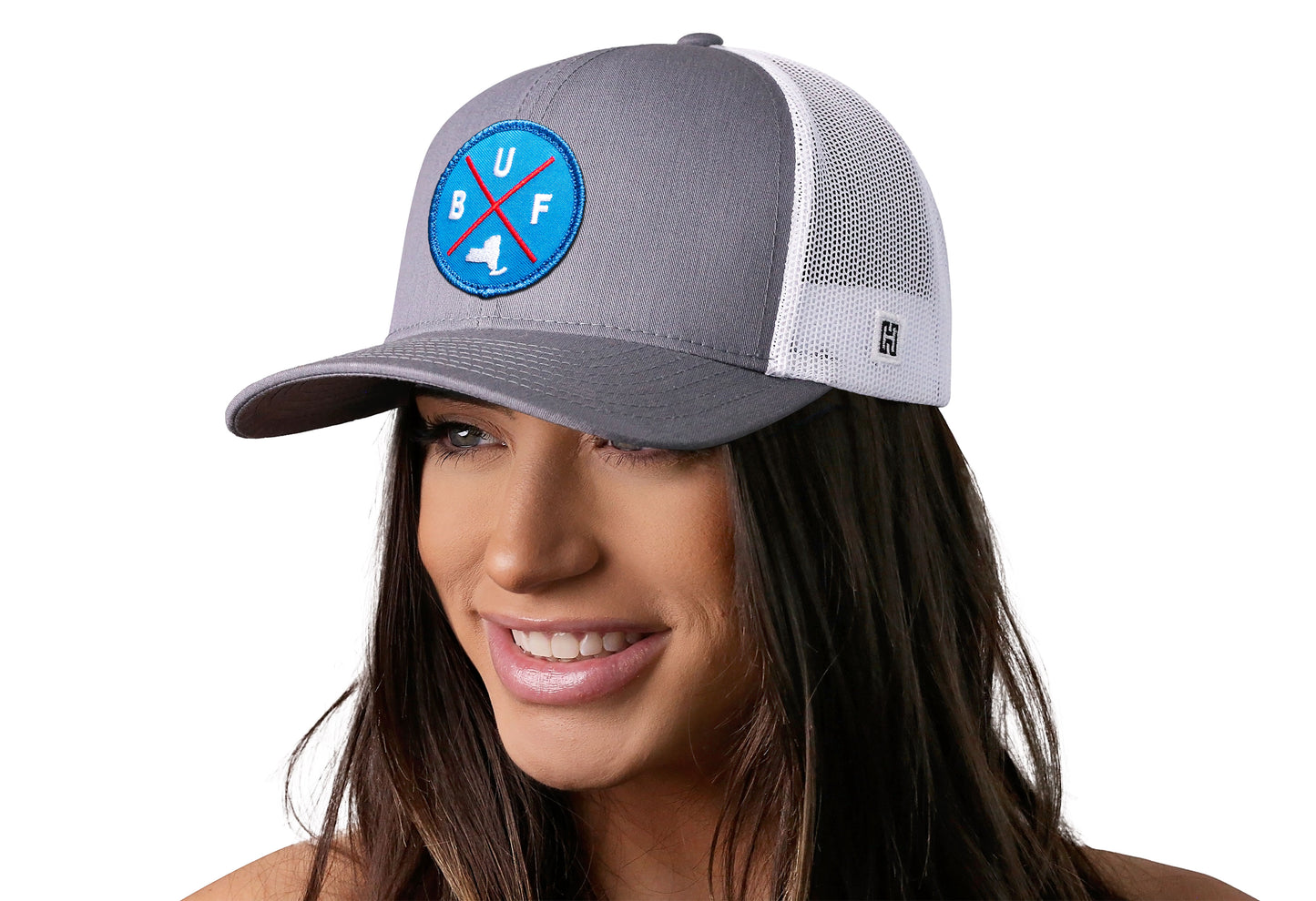 Gray-White trucker hat worn by a female model viewed from the left side and features a Buffalo BUF X Red, White, Blue Embroidered Circle Patch displayed on the front panel. The hat has a classic mesh back design and a curved structured bill