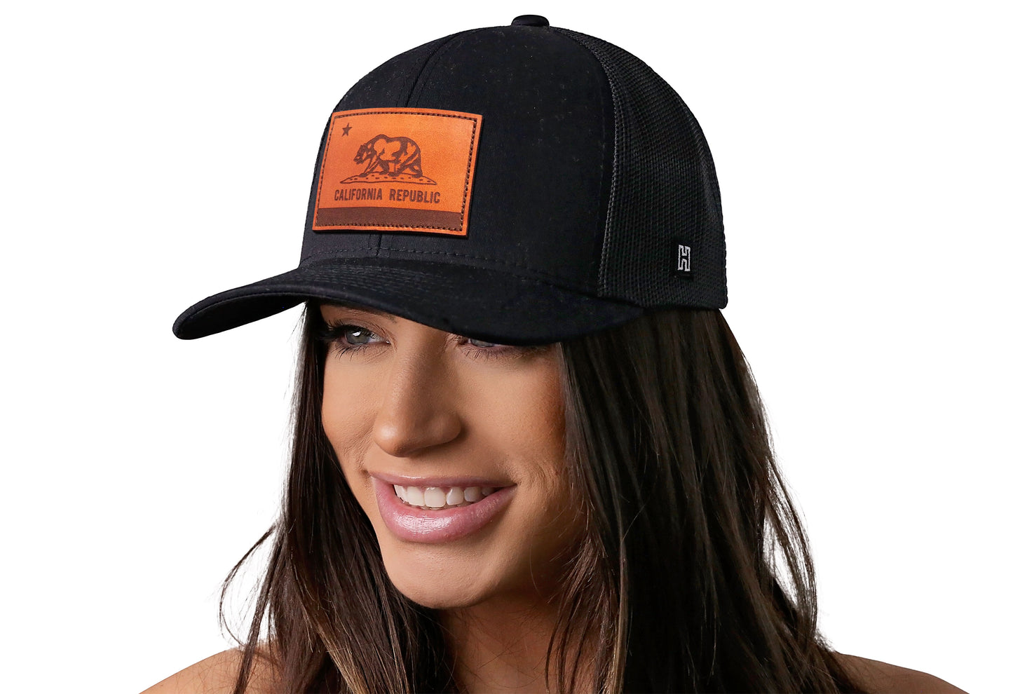 Black trucker hat worn by a female model viewed from the left side and features a California Flag Leather Rectangle Patch displayed on the front panel. The hat has a classic mesh back design and a curved structured bill