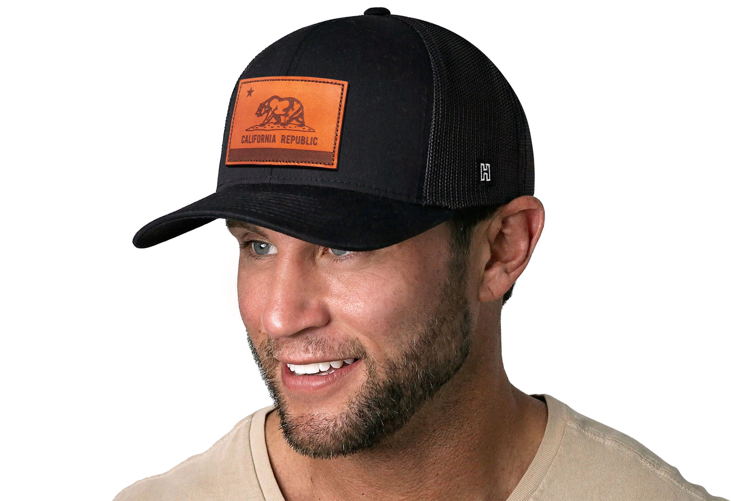 Black trucker hat worn by a male model viewed from the left side and features a California Flag Leather Rectangle Patch displayed on the front panel. The hat has a classic mesh back design and a curved structured bill