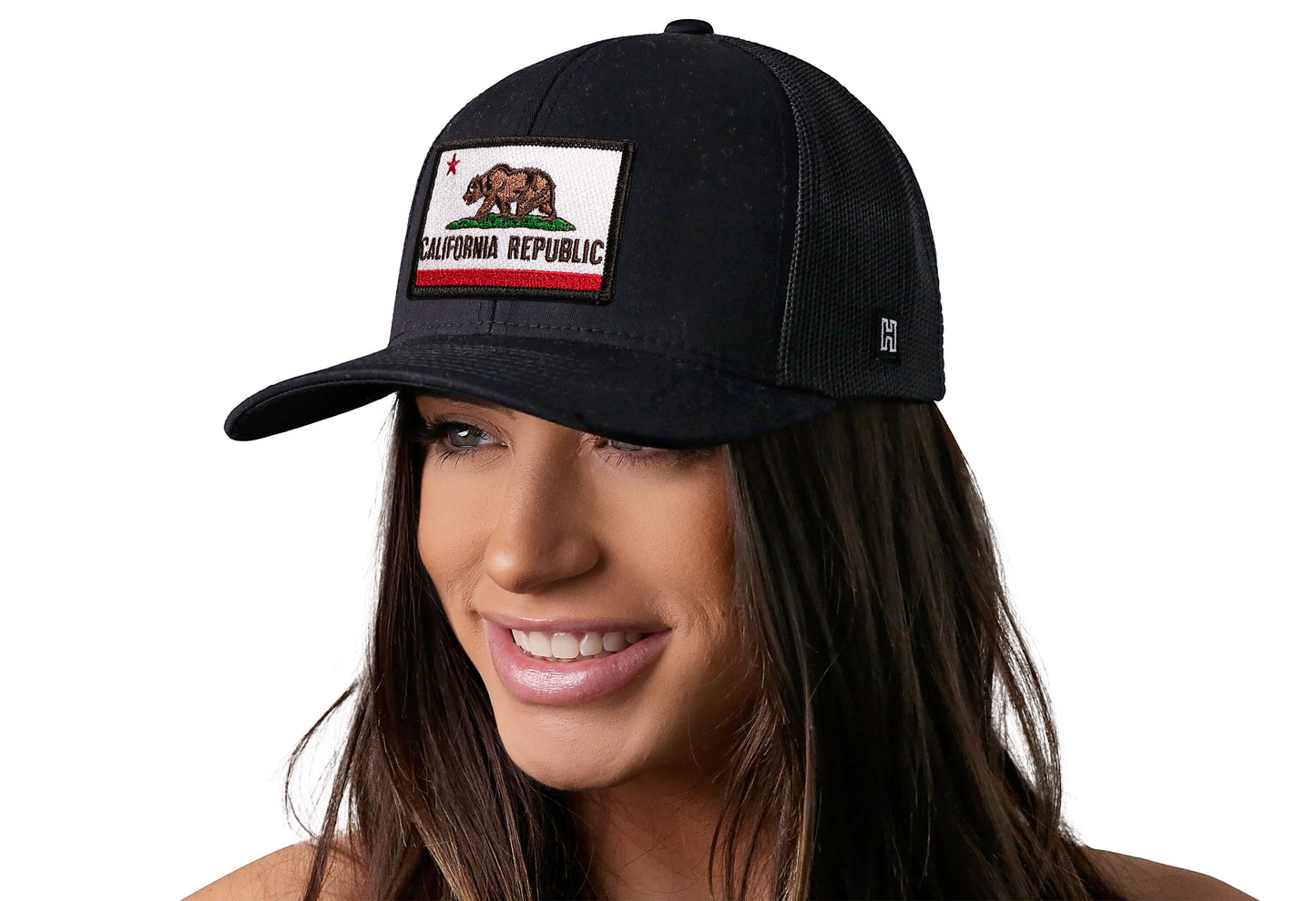 Black trucker hat worn by a female model viewed from the left side and features a California Flag Embroidered Rectangle Patch displayed on the front panel. The hat has a classic mesh back design and a curved structured bill