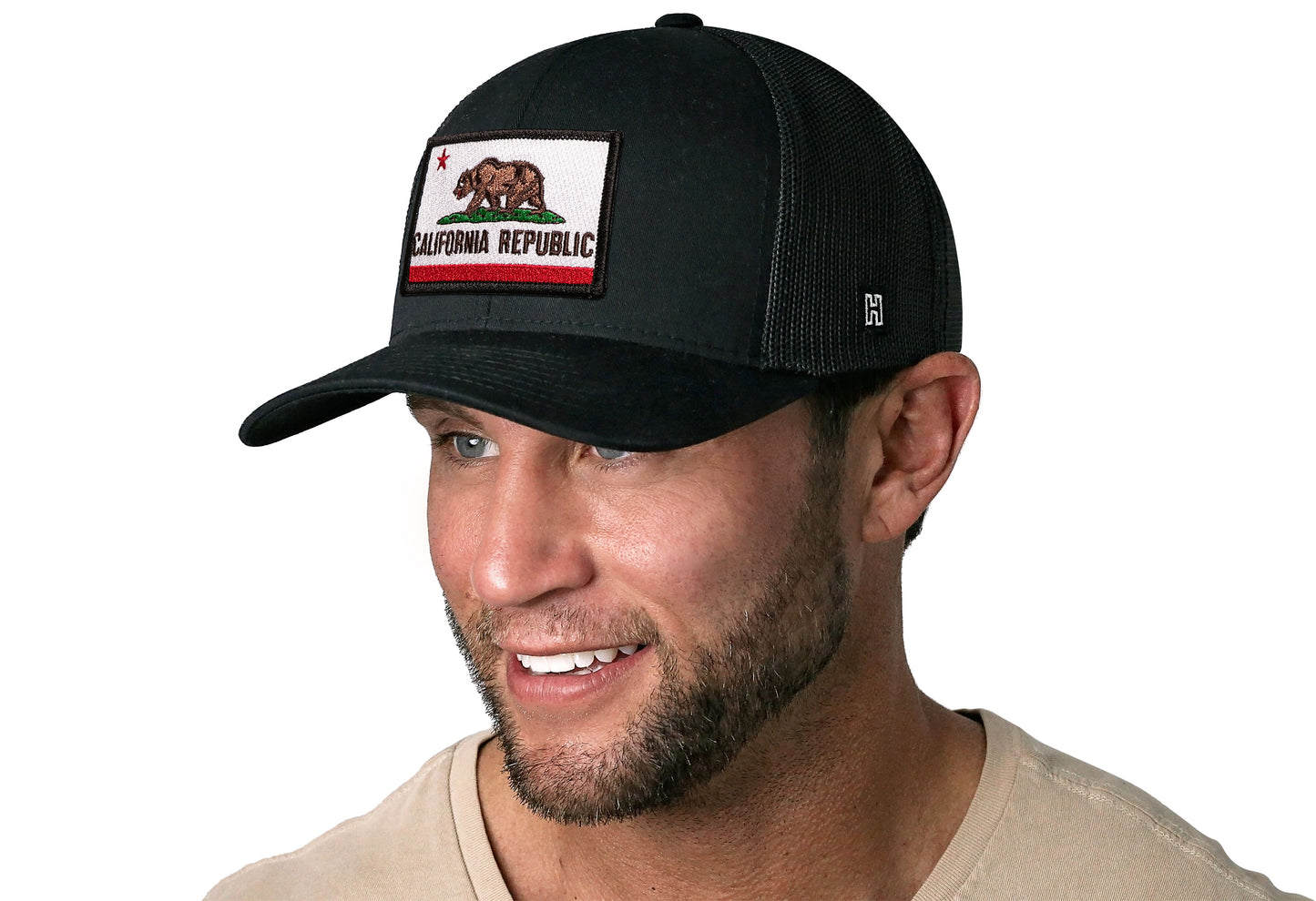 Black trucker hat worn by a male model viewed from the left side and features a California Flag Embroidered Rectangle Patch displayed on the front panel. The hat has a classic mesh back design and a curved structured bill